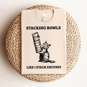 Könnte beinhalten: Ein beiges T-Shirt mit einer schwarz-weißen Illustration einer Katze, die einen Stapel Schalen hält. Der Text "Stacking Bowls Like I Stack Excuses" ist auf das T-Shirt gedruckt.