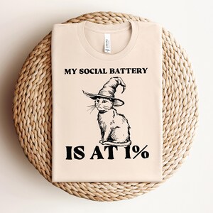 Op de afbeelding: Een beige T-shirt met de tekst "My social battery is at 1%" en een zwart-witte illustratie van een kat met een heksenhoed.