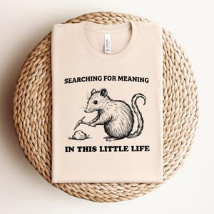 May include: A beige t-shirt with the text "SEARCHING FOR MEANING IN THIS LITTLE LIFE" and a black and white illustration of a mouse digging with a pickaxe. The shirt is displayed on a woven, circular placemat.