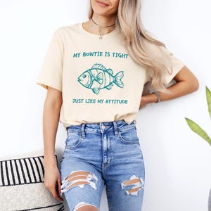 May include: A light beige t-shirt with a teal blue fish graphic and the text "My Bowtie is Tight Just Like My Attitude".