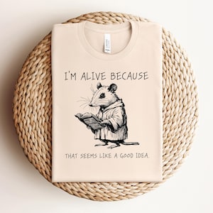 Może przedstawiać: Beżowa koszulka z czarno-białym rysunkiem myszy czytającej książkę. Tekst na koszulce brzmi: "Jestem żywy, bo to wydaje się dobrym pomysłem."