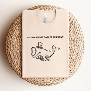 May include: A light beige t-shirt with a black whale wearing a top hat and the text "Ocean's Most Dapper Resident".