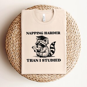 May include: A beige t-shirt with black text that reads "NAPPING HARDER THAN I STUDIED". A cartoon raccoon wearing a graduation cap and holding a scroll is pictured below the text.