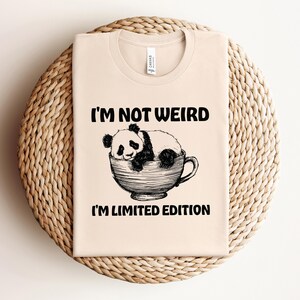 Puede incluir: Una camiseta beige con una ilustración en blanco y negro de un panda en una taza de té. El texto "I'm not weird I'm limited edition" está impreso en la camiseta.