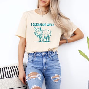 Op de afbeelding: Een licht beige t-shirt met een afbeelding van een varken met een strik en de tekst "I clean up well".