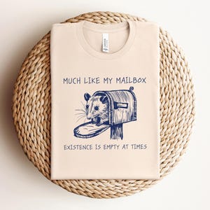 Puede incluir: Una camiseta beige con una ilustración azul de una zarigüeya mirando dentro de un buzón. El texto "Much like my mailbox, existence is empty at times" está impreso en la camiseta.
