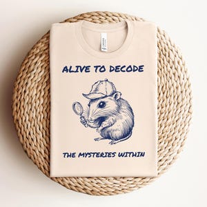 Puede incluir: Una camiseta beige con una ilustración azul de un ratón con un sombrero de detective y sosteniendo una lupa. El texto "Alive to Decode the Mysteries Within" está impreso en la camiseta.