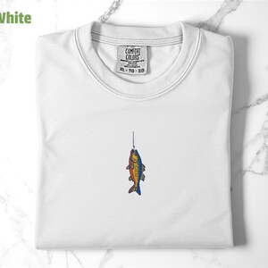 May include: White t-shirt with a colourful fish graphic on the front. The fish is on a fishing line and has a red, orange, yellow, blue, and green colour scheme. The shirt has a tag that says "COMFORT COLORS" and "XL - TG - EG".
