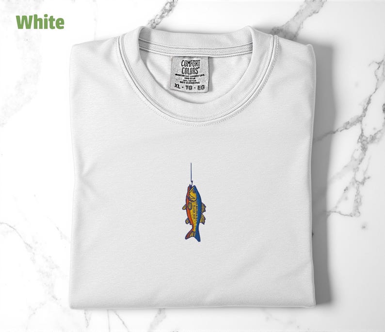 May include: White t-shirt with a colourful fish graphic on the front. The fish is on a fishing line and has a red, orange, yellow, blue, and green colour scheme. The shirt has a tag that says "COMFORT COLORS" and "XL - TG - EG".