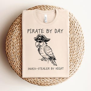 Puede incluir: Una camiseta beige con una ilustración en blanco y negro de un loro con un sombrero de pirata. El texto "Pirate by Day Snack-Stealer by Night" está impreso en la camiseta.