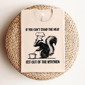 May include: A beige t-shirt with a black and white illustration of a skunk wearing a chef's hat and holding a spoon over a pot. The text "If you can't stand the heat get out of the kitchen" is printed on the shirt.