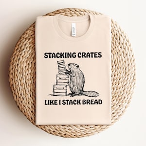 Puede incluir: Una camiseta beige con un gráfico negro de un castor apilando cajas. El texto "Stacking Crates Like I Stack Bread" está impreso debajo del gráfico.