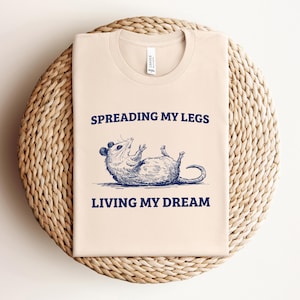 Puede incluir: Una camiseta beige con una ilustración azul de un zarigüeya extendiendo sus piernas y el texto "Spreading my legs living my dream".