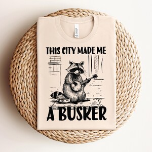 Può includere: Una maglietta beige con un'illustrazione in bianco e nero di un procione che suona il banjo. Il testo "This city made me a busker" è stampato sulla maglietta.
