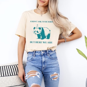 May include: A cream colored t-shirt with a panda wearing a party hat and the text "I didn't ask to be born but here we are".