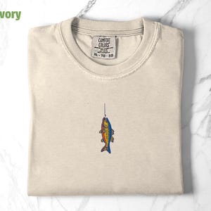 May include: A beige t-shirt with a small embroidered image of a fish on a hook. The t-shirt has a tag that says "COMFORT COLORS" and "XL - TG - EG".