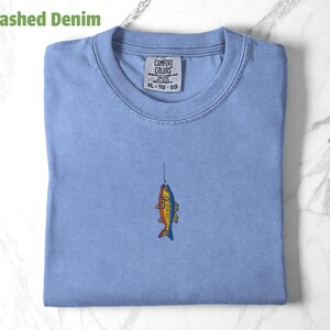 May include: A light blue t-shirt with a small embroidered image of a fish on a fishing line. The shirt has a tag that reads "Comfort Colors" and "XL - TG - EG".
