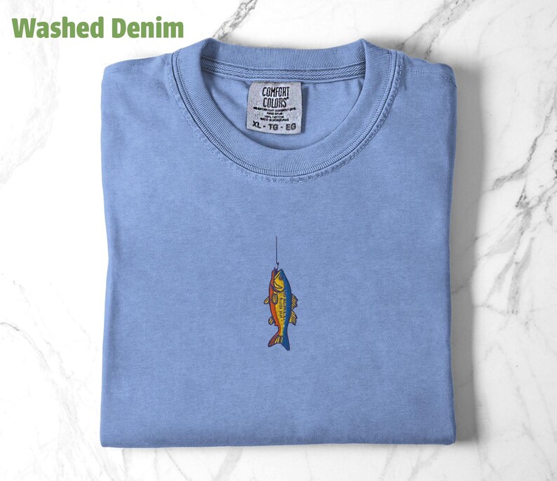 May include: A light blue t-shirt with a small embroidered image of a fish on a fishing line. The shirt has a tag that reads "Comfort Colors" and "XL - TG - EG".