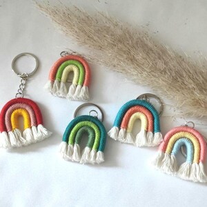 Könnte beinhalten: Fünf regenbogenfarbene gehäkelte Schlüsselanhänger mit weißen Quasten. Jeder Schlüsselanhänger hat eine andere Farbkombination aus Rot, Orange, Gelb, Grün, Blau und Lila.