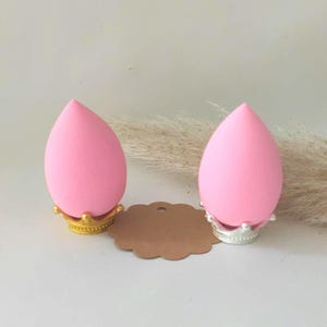 Könnte beinhalten: Zwei rosa Make-up-Schwämme in Tropfenform, die jeweils auf einem dekorativen kronenförmigen Ständer stehen. Ein Ständer ist golden, der andere ist silber.