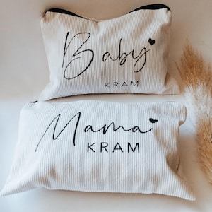 Könnte beinhalten: Zwei cremefarbene Cordkissen mit schwarzem Besatz. Das obere Kissen trägt das Wort "Baby" und das untere Kissen das Wort "Mama" in schwarzer Schrift. Beide Kissen haben das Wort "KRAM" darunter.