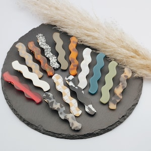 Könnte beinhalten: Eine Sammlung von wellenförmigen Haarspangen in verschiedenen Farben und Mustern, darunter Rot, Weiß, Schildpatt und gesprenkelte Designs. Die Spangen sind auf einer dunkelgrauen, runden, strukturierten Oberfläche angeordnet, mit neutralem Hintergrund und getrocknetem Ziergras.