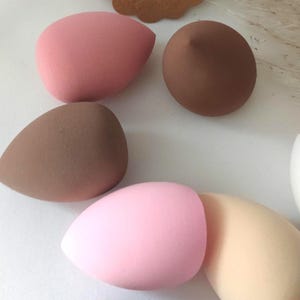 Könnte beinhalten: Ein Set mit sechs Make-up-Schwämmen in verschiedenen Farben, darunter Rosa, Braun und Beige. Die Schwämme sind tropfenförmig und zum Verblenden von Make-up gedacht.