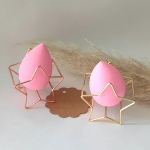 Könnte beinhalten: Zwei rosa Make-up-Schwämme werden auf goldenen sternförmigen Ständern präsentiert. Die Ständer bestehen aus dünnem Metalldraht und haben ein geometrisches Design.