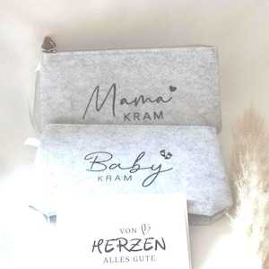 Könnte beinhalten: Zwei hellgraue Filztaschen mit schwarzer Schrift, die "Mama KRAM" und "Baby KRAM" lautet. Vor den Taschen liegt eine weiße Karte mit schwarzem Text "VON HERZEN ALLES GUTE". Die Taschen haben einen Reißverschluss und einen kleinen braunen Zipper.