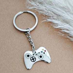 Könnte beinhalten: Ein Schlüsselanhänger aus silberfarbenem Metall mit einem Anhänger in Form eines Gamecontrollers. Der Anhänger hat eine glatte, reflektierende Oberfläche mit schwarzen kreisförmigen Details, die Joysticks und Tasten darstellen, sowie vier kleinen, farbigen Emaille-Punkten in Rot, Gelb, Grün und Blau.