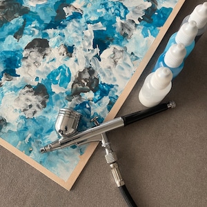 Könnte beinhalten: Ein silberner Airbrush mit schwarzem Griff und silberner Düse. Der Airbrush liegt auf einer weißen Oberfläche mit einem blauen und weißen abstrakten Gemälde im Hintergrund. Im Hintergrund befinden sich mehrere kleine blaue Flaschen mit weißen Kappen.
