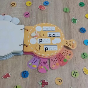 May include: A colorful alphabet learning book with a yellow cover and a cut-out shape of a hand. The book has a page with the letter 'P' and a page with the alphabet in a rainbow pattern. There are also colorful letter stickers on a wooden surface.