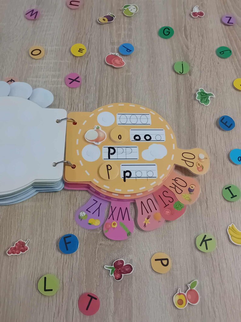 May include: A colorful alphabet learning book with a yellow cover and a cut-out shape of a hand. The book has a page with the letter 'P' and a page with the alphabet in a rainbow pattern. There are also colorful letter stickers on a wooden surface.