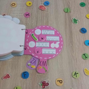 May include: Pink alphabet learning toy with a book and letter tiles. The book has pages with letters and lines for tracing. The tiles are colorful and have letters on them.