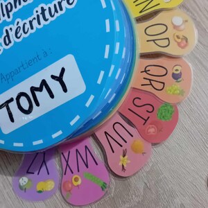 May include: A colorful alphabet learning toy with a blue circle that says "alphabet d'&eacute;criture" and "Appartient &agrave;: TOMY". The toy has colorful tabs with letters and pictures of fruits and vegetables.