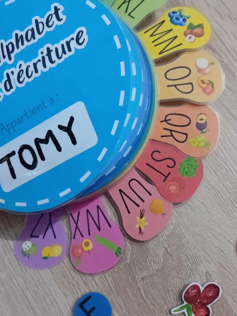 May include: A colorful alphabet learning toy with a blue circle that says "alphabet d'&eacute;criture" and "Appartient &agrave;: TOMY". The toy has colorful tabs with letters and pictures of fruits and vegetables.
