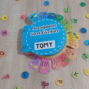 May include: A blue and rainbow alphabet book with the title "Mon alphabet Livret d'&eacute;criture" and a personalized name tag that says "TOMY". The book is open to reveal the letters of the alphabet with colorful illustrations of fruits and vegetables.
