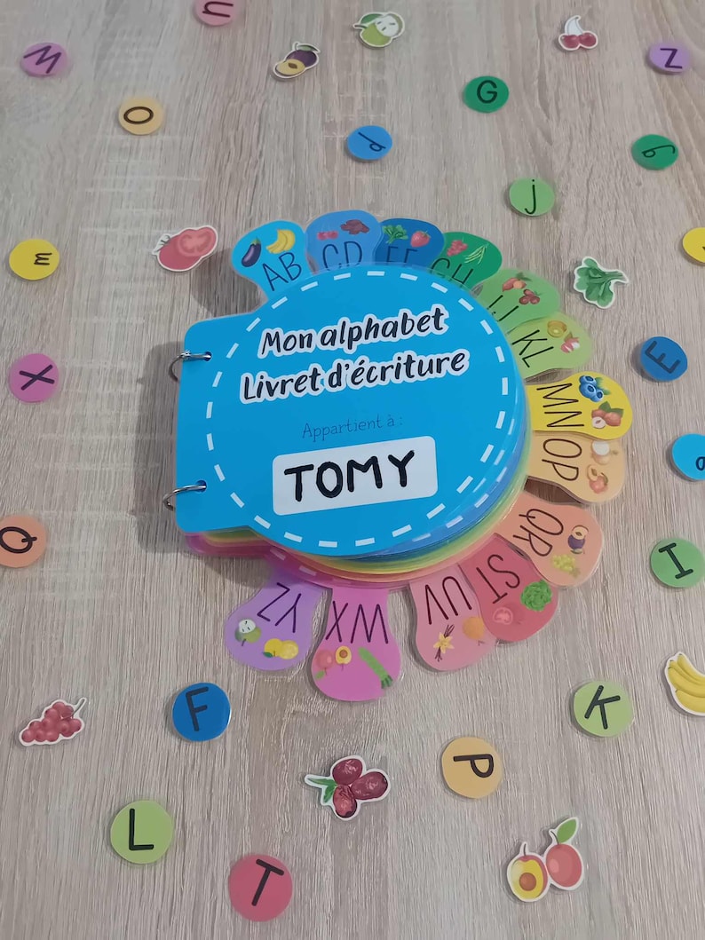 May include: A blue and rainbow alphabet book with the title "Mon alphabet Livret d'&eacute;criture" and a personalized name tag that says "TOMY". The book is open to reveal the letters of the alphabet with colorful illustrations of fruits and vegetables.