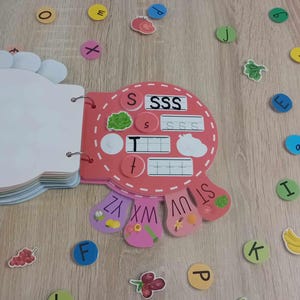 May include: A pink and red alphabet learning toy with a flap book and a spinning wheel with letters S, T, U, V, W, X, Y, and Z. The toy has a section for learning the letter S with the words "SSS" and "SSSS" printed on it.