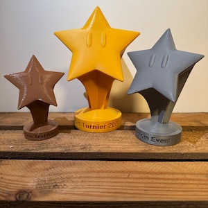 Könnte beinhalten: Drei 3D-gedruckte sternförmige Trophäen mit einem Comic-Gesicht auf jeder. Die größte Trophäe ist gelb, die mittlere Trophäe ist braun und die kleinste Trophäe ist grau. Die Basis jeder Trophäe hat Text darauf. Die größte Trophäe sagt "Kart Turnier 2023", die mittlere Trophäe sagt "Dein Text hier" und die kleinste Trophäe sagt "Custom Event".