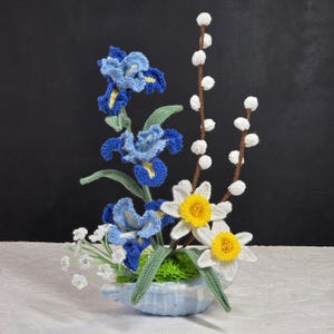May include: A crocheted floral arrangement in a blue and white ceramic vase. The arrangement features blue and yellow irises, white daffodils, and white pussy willow branches.