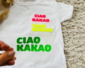 Può includere: Una maglietta bianca con le parole "CIAO KAKAO" in rosso, giallo e verde. La maglietta è esposta su una superficie strutturata. Il testo è in un carattere giocoso.