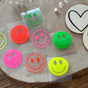 Könnte beinhalten: Eine Auswahl an bunten Smiley-Aufnähern in Neonfarben wie Gelb, Orange, Pink und Grün. Zwei Holzuntersetzer mit Herzmotiven sind ebenfalls zu sehen, einer mit schwarzem Herz und einer mit schwarzer Umrandung.