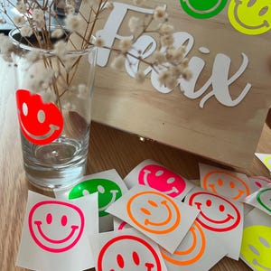 Könnte beinhalten: Eine Holzkiste mit dem Namen "Felix" in weißer Schrift, verziert mit grünen und gelben Smiley-Aufklebern. Ein klares Glas mit einem roten Smiley-Design und mehrere Smiley-Aufkleber in verschiedenen Farben sind ebenfalls zu sehen.