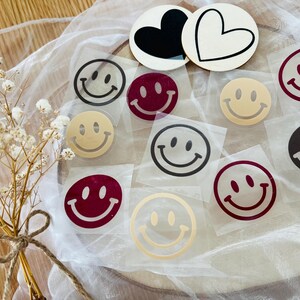 Könnte beinhalten: Eine Sammlung von Smiley-Aufklebern in verschiedenen Farben, darunter Schwarz, Burgunderrot und Gold. Einige Aufkleber befinden sich auf transparentem Träger, andere auf herzförmigen Holzzuschnitten. Ein kleiner Strauß Trockenblumen befindet sich im Vordergrund.