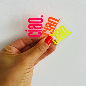 Könnte beinhalten: Drei transparente Aufkleber mit dem Wort "ciao" in leuchtenden Neonfarben: Pink, Orange und Gelb. Die Aufkleber werden vor einem weißen Hintergrund gehalten. Das Wort "ciao" ist in einer fetten serifenlosen Schrift geschrieben.