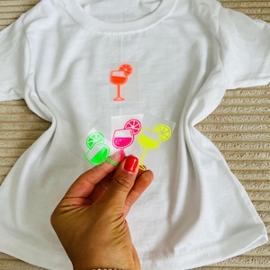Könnte beinhalten: Weißes T-Shirt mit einer neongelben Cocktailgrafik. Eine Hand hält drei zusätzliche Cocktailgrafiken in Neon-Grün, Pink und Gelb. Jede Grafik zeigt ein Cocktailglas mit einer Zitrusfruchtscheibe. Das Shirt liegt auf einer beigen Oberfläche.