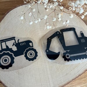 Könnte beinhalten: Zwei schwarze Silhouette-Aufkleber von Baumaschinen. Einer ist ein Traktor und der andere ein Bagger. Die Aufkleber befinden sich auf einer hellbraunen Holzoberfläche.