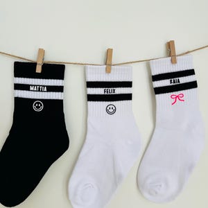 Könnte beinhalten: Drei Socken hängen an einer Schnur mit Holzklammern. Die erste ist schwarz mit weißen Streifen und dem Namen MATTIA über einem Smiley. Die zweite ist weiß mit schwarzen Streifen und dem Namen FELIX über einem Smiley. Die dritte ist weiß mit schwarzen Streifen und dem Namen KAIA über einer rosa Schleife.