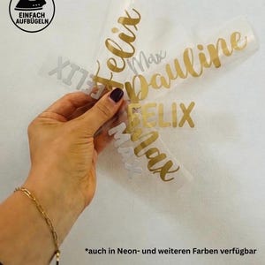 Könnte beinhalten: Goldene und silberne Wärmeübertragungs-Vinylbuchstaben, die Namen wie "Felix", "Max" und "Pauline" buchstabieren. Das Bild enthält ein Bügelsymbol und den Text "Einfach Aufbügeln". Der Text "*auch in Neon- und weiteren Farben verfügbar" ist ebenfalls sichtbar.